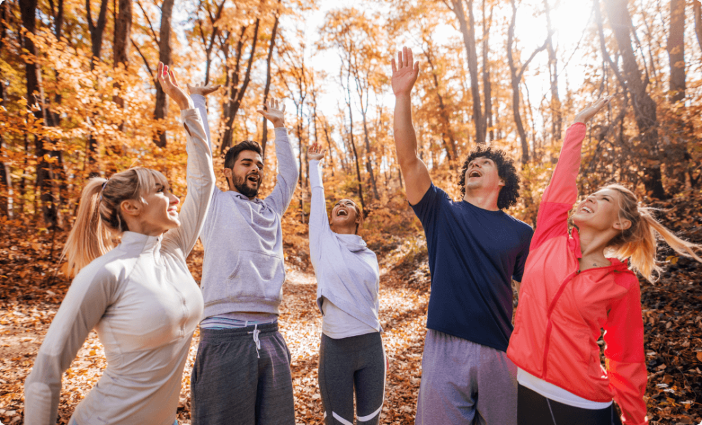 A group of young men and women having fun and raising their hands while walking on a forest trail among fall leaves and autumnal colors.